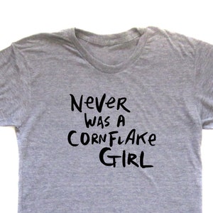 May include: A heather gray t-shirt with the text "Never Was a Cornflake Girl" in black, handwritten-style lettering. The shirt is a basic crew neck style.