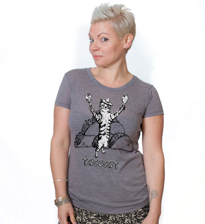 May include: A heather grey t-shirt with a black and white illustration of a cat wearing a sombrero and holding maracas. The cat is standing on top of two tacos. The text "tacocat" is printed below the cat.