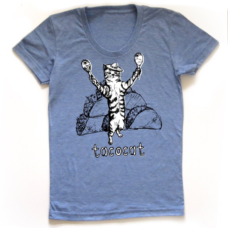May include: A light blue t-shirt with a black and white illustration of a cat wearing a sombrero and holding maracas. The cat is standing on top of a pile of tacos. The text "tacocat" is printed below the cat.