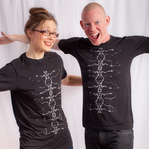 May include: Two people wearing black t-shirts with a white graphic design of a spine. The person on the left is a woman with brown hair and glasses. The person on the right is a man with short, shaved hair. Both are smiling.