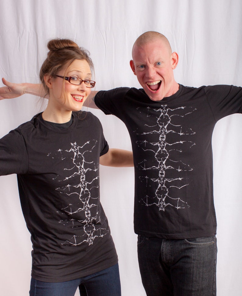 May include: Two people wearing black t-shirts with a white graphic design of a spine. The person on the left is a woman with brown hair and glasses. The person on the right is a man with short, shaved hair. Both are smiling.