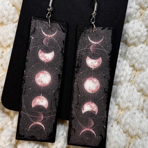 Può includere: Un paio di orecchini rettangolari con un design celeste. Ogni orecchino presenta uno sfondo nero con un'illustrazione delle fasi lunari rosa. Gli orecchini hanno ganci color argento.