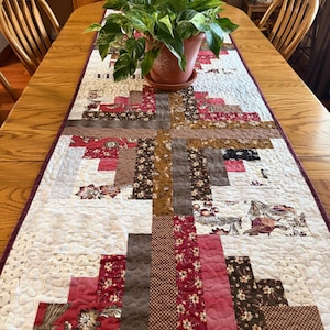 May include: A quilted table runner with a patchwork design in shades of red, brown, and cream. The runner features floral and geometric patterns, with a dark purple border. A potted plant sits on the runner.