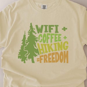 A light yellow t-shirt with the words "WIFI + COFFEE + HIKING = FREEDOM" in green and orange lettering. Two green tree graphics are on the left side of the shirt. The shirt is made of a soft material.