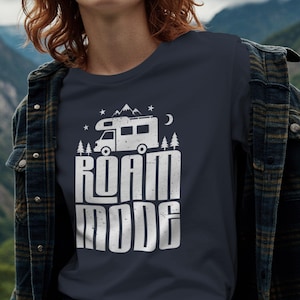 May include: A dark charcoal gray t-shirt featuring a white graphic print of a camper van, pine trees, mountains, and stars above the text "ROAM MODE" in a bold, distressed font. The shirt is worn under a dark plaid button-down flannel shirt.