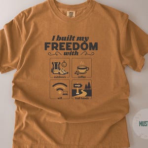 Mustard-colored t-shirt with the text "I built my freedom with" and icons representing outdoors, coffee, wifi, and trail heads. The shirt has a crew neck and short sleeves.