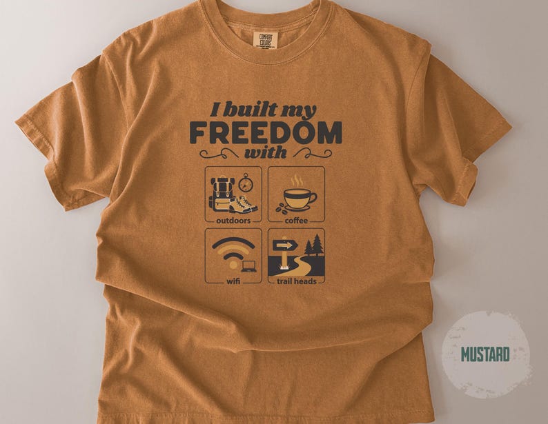 Mustard-colored t-shirt with the text "I built my freedom with" and icons representing outdoors, coffee, wifi, and trail heads. The shirt has a crew neck and short sleeves.