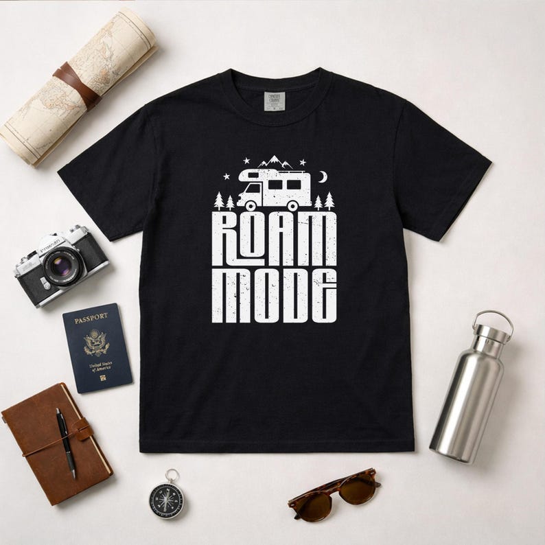 May include: A black short-sleeved t-shirt featuring a white graphic print of a camper van, pine trees, mountains, stars, and a crescent moon above the text ROAM MODE in a distressed, bold, sans-serif font centered on the chest area.