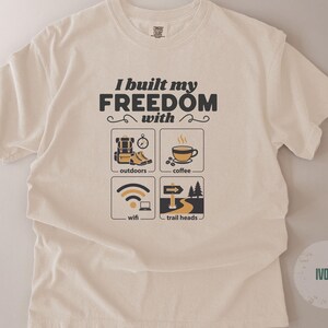 Ivory-colored t-shirt with the text "I built my freedom with" above four small illustrations. The illustrations depict outdoor gear, coffee, wifi, and trailheads. The shirt is a crew neck.