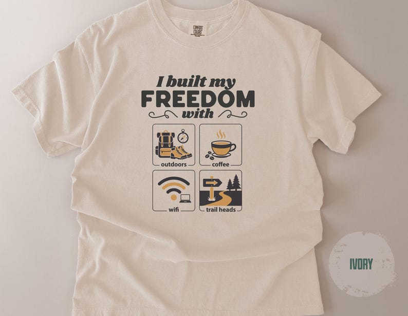 Ivory-colored t-shirt with the text "I built my freedom with" above four small illustrations. The illustrations depict outdoor gear, coffee, wifi, and trailheads. The shirt is a crew neck.