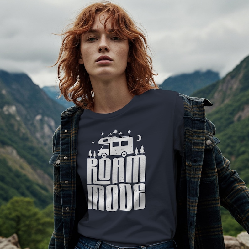 May include: A dark charcoal gray t-shirt featuring a white graphic print of a camper van, pine trees, mountains, and stars above the text "ROAM MODE" in a bold, distressed font. The shirt is worn under a dark plaid button-down flannel shirt.