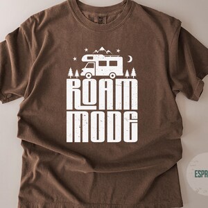 May include: A brown short-sleeve t-shirt featuring a white graphic print of a camper van, mountains, trees, stars, and a crescent moon. Below the graphic, the text reads ROAM MODE in a bold, distressed white font. The shirt has a crew neckline.