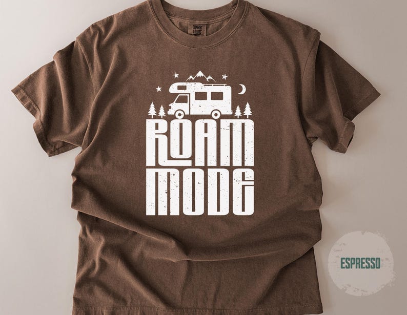 May include: A brown short-sleeve t-shirt featuring a white graphic print of a camper van, mountains, trees, stars, and a crescent moon. Below the graphic, the text reads ROAM MODE in a bold, distressed white font. The shirt has a crew neckline.