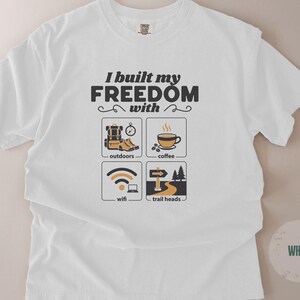 White t-shirt with the text "I built my FREEDOM with" above four icons: outdoors, coffee, wifi, and trail heads. The shirt has a crew neck and short sleeves. The design is in black and tan.