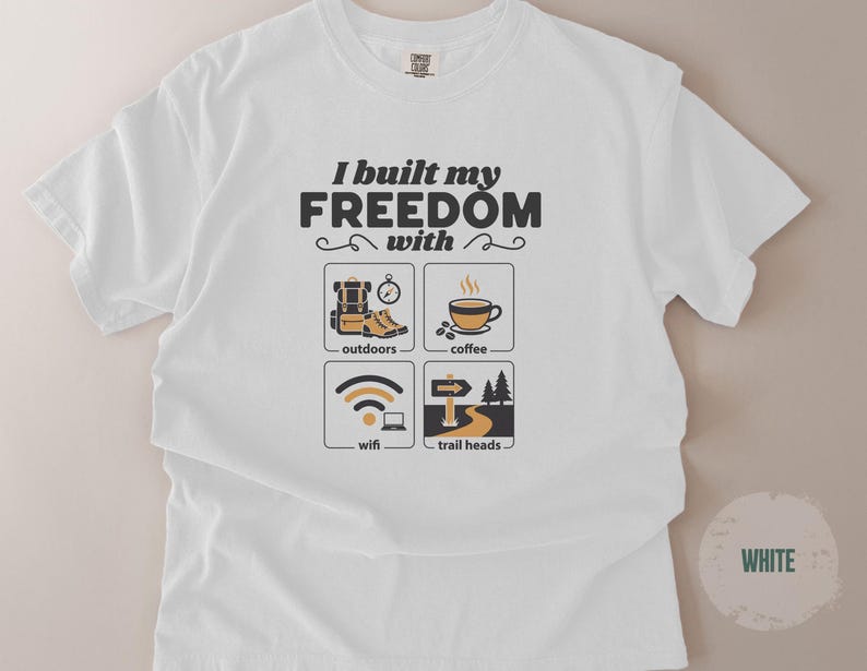 White t-shirt with the text "I built my FREEDOM with" above four icons: outdoors, coffee, wifi, and trail heads. The shirt has a crew neck and short sleeves. The design is in black and tan.