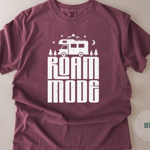 May include: A berry-colored short-sleeve t-shirt featuring a white graphic print of a camper van, pine trees, mountains, stars, and a crescent moon. Below the graphic, the text reads ROAM MODE in a bold, distressed, white sans-serif font.