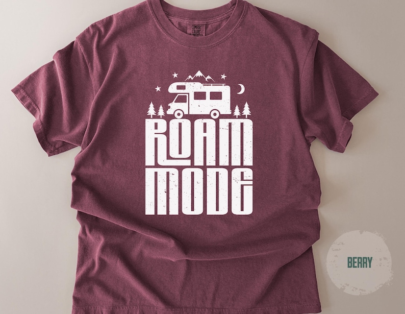 May include: A berry-colored short-sleeve t-shirt featuring a white graphic print of a camper van, pine trees, mountains, stars, and a crescent moon. Below the graphic, the text reads ROAM MODE in a bold, distressed, white sans-serif font.