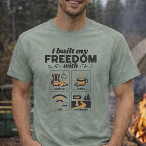 Sage green t-shirt with the text "I built my FREEDOM with" and icons representing outdoors, coffee, wifi, and trail heads. The shirt is a crew neck and has short sleeves.