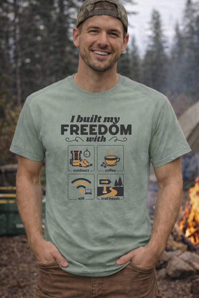 Sage green t-shirt with the text "I built my FREEDOM with" and icons representing outdoors, coffee, wifi, and trail heads. The shirt is a crew neck and has short sleeves.