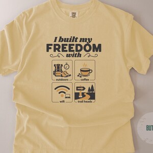 Butter-colored t-shirt with the text "I built my FREEDOM with" above four illustrated icons: outdoors, coffee, wifi, and trail heads. The shirt has a crew neck and short sleeves.