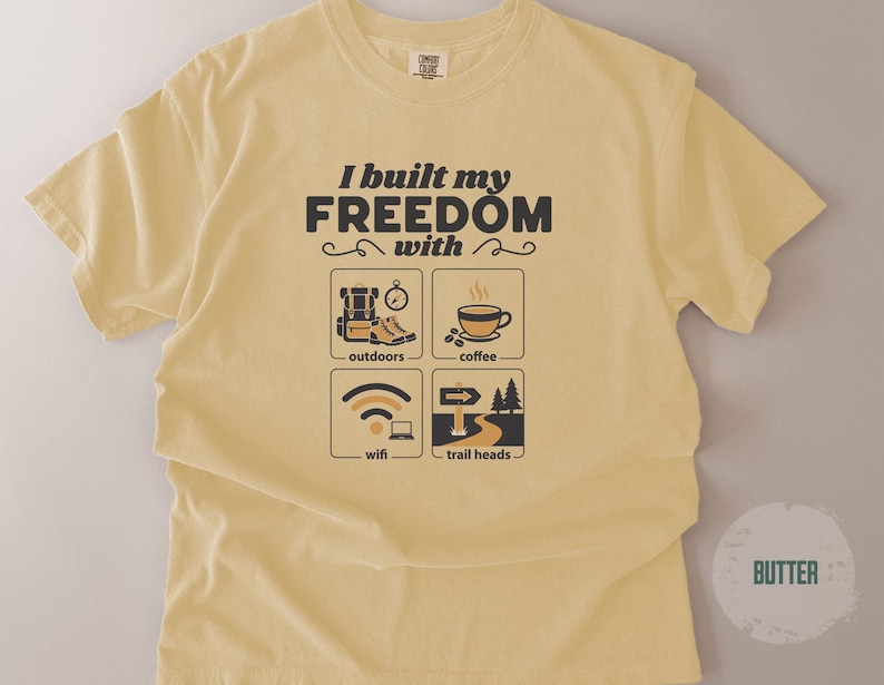 Butter-colored t-shirt with the text "I built my FREEDOM with" above four illustrated icons: outdoors, coffee, wifi, and trail heads. The shirt has a crew neck and short sleeves.