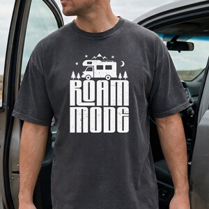 May include: A charcoal gray short-sleeve t-shirt featuring a white graphic print of a camper van, pine trees, mountains, stars, and a crescent moon above the text ROAM MODE in a bold, distressed, block-style font centered on the chest.