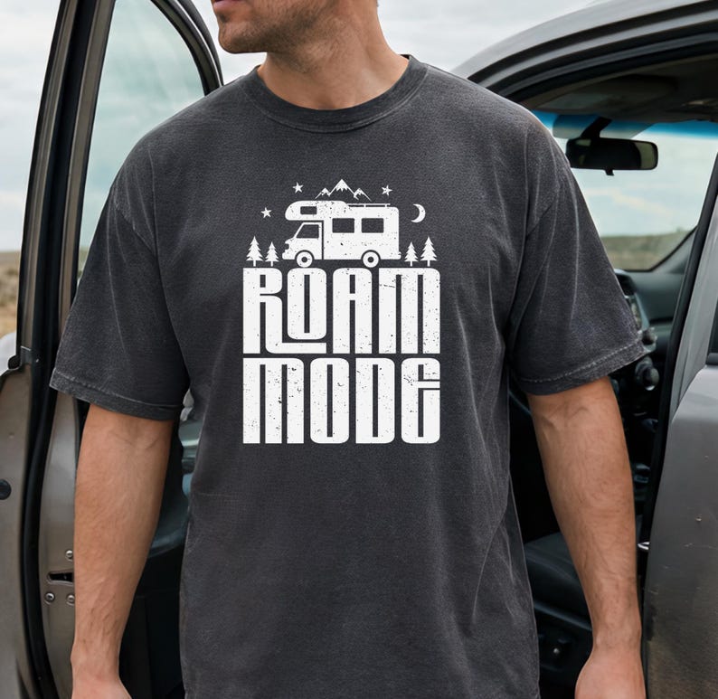May include: A charcoal gray short-sleeve t-shirt featuring a white graphic print of a camper van, pine trees, mountains, stars, and a crescent moon above the text ROAM MODE in a bold, distressed, block-style font centered on the chest.