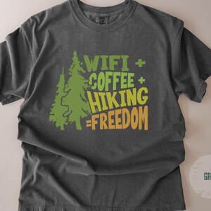 Dark grey t-shirt with the text "WIFI + COFFEE + HIKING = FREEDOM" in green and orange. Two green pine trees are on the left side of the text. The shirt is a comfortable, casual style.