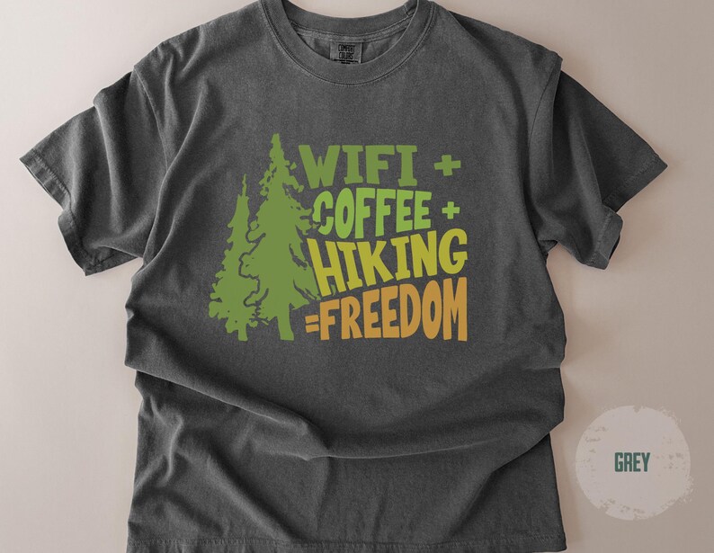 Dark grey t-shirt with the text "WIFI + COFFEE + HIKING = FREEDOM" in green and orange. Two green pine trees are on the left side of the text. The shirt is a comfortable, casual style.