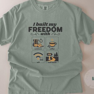 Sage green t-shirt with the text "I built my FREEDOM with" and icons representing outdoors, coffee, wifi, and trailheads. The shirt has a crew neck and short sleeves. The word "BAY" is printed on a circle in the lower right corner.