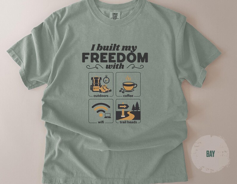 Sage green t-shirt with the text "I built my FREEDOM with" and icons representing outdoors, coffee, wifi, and trailheads. The shirt has a crew neck and short sleeves. The word "BAY" is printed on a circle in the lower right corner.