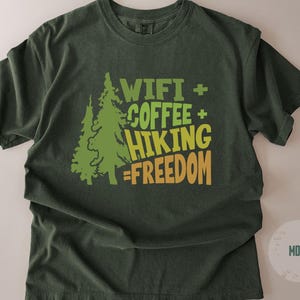 A moss green t-shirt with a graphic design. The design includes two green pine trees and the words "WIFI + COFFEE + HIKING = FREEDOM" in green and yellow lettering. The shirt is a casual style.