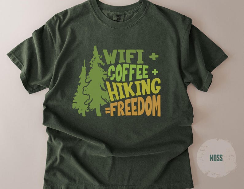 A moss green t-shirt with a graphic design. The design includes two green pine trees and the words "WIFI + COFFEE + HIKING = FREEDOM" in green and yellow lettering. The shirt is a casual style.