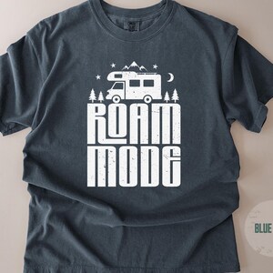 May include: A dark blue short-sleeve t-shirt featuring a white graphic print of a camper van, pine trees, mountains, stars, and a crescent moon. The text ROAM MODE is printed in large, bold, distressed white capital letters below the camper van.