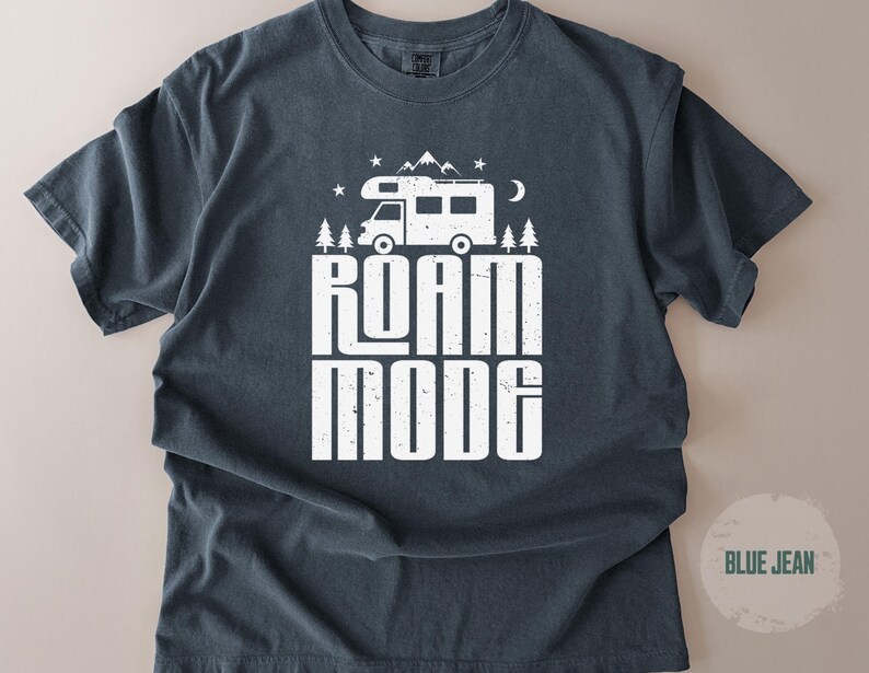 May include: A dark blue short-sleeve t-shirt featuring a white graphic print of a camper van, pine trees, mountains, stars, and a crescent moon. The text ROAM MODE is printed in large, bold, distressed white capital letters below the camper van.