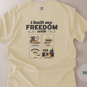 A light yellow t-shirt with the text "I built my FREEDOM with" above four illustrated icons: outdoors, coffee, wifi, and trail heads. The shirt is a soft, comfortable material.