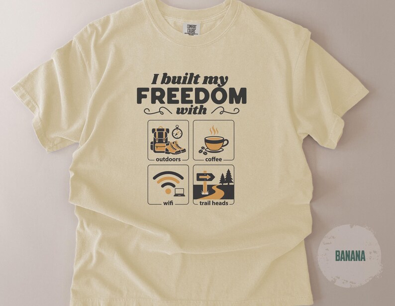 A light yellow t-shirt with the text "I built my FREEDOM with" above four illustrated icons: outdoors, coffee, wifi, and trail heads. The shirt is a soft, comfortable material.