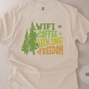 Ivory-colored t-shirt with a crew neck. The front features a green tree graphic and the words "WIFI + COFFEE + HIKING = FREEDOM" in green and yellow-orange lettering. The shirt is made of a soft, comfortable material.