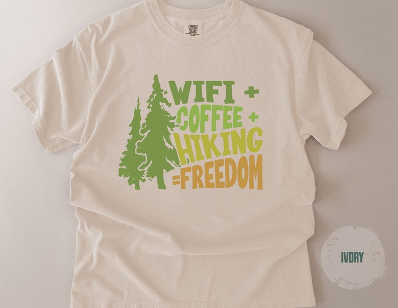 Ivory-colored t-shirt with a crew neck. The front features a green tree graphic and the words "WIFI + COFFEE + HIKING = FREEDOM" in green and yellow-orange lettering. The shirt is made of a soft, comfortable material.