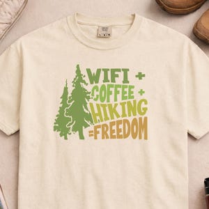 Cream-colored t-shirt with a green and brown graphic. The graphic includes two stylized trees and the words "WIFI + COFFEE + HIKING = FREEDOM" in green and brown. The shirt is made of a soft material.
