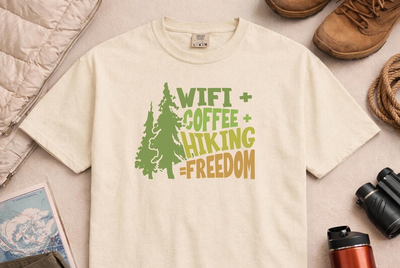 Cream-colored t-shirt with a green and brown graphic. The graphic includes two stylized trees and the words "WIFI + COFFEE + HIKING = FREEDOM" in green and brown. The shirt is made of a soft material.
