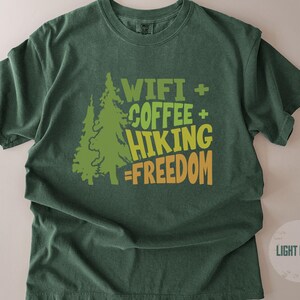 A light green t-shirt with the text "WIFI + COFFEE + HIKING = FREEDOM" in green and orange. Two green pine trees are to the left of the text. The shirt is a comfortable, casual style.