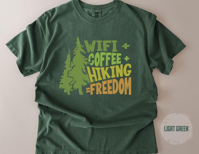 A light green t-shirt with the text "WIFI + COFFEE + HIKING = FREEDOM" in green and orange. Two green pine trees are to the left of the text. The shirt is a comfortable, casual style.