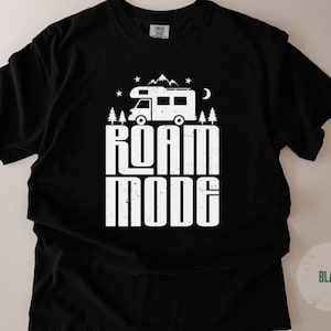 May include: A black short-sleeve t-shirt featuring a white graphic print of a camper van, pine trees, mountains, stars, and a crescent moon. The text ROAM MODE is printed in large, bold, distressed white capital letters below the camper van graphic.