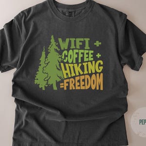 A dark gray t-shirt with the words "WIFI + COFFEE + HIKING = FREEDOM" in green and orange lettering. Two green pine trees are on the left side of the text. The shirt is made of a soft material.