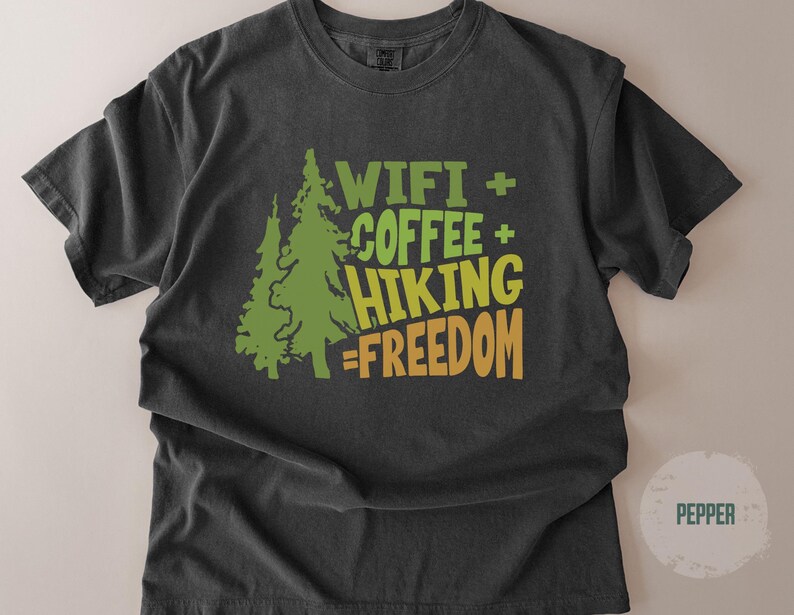 A dark gray t-shirt with the words "WIFI + COFFEE + HIKING = FREEDOM" in green and orange lettering. Two green pine trees are on the left side of the text. The shirt is made of a soft material.