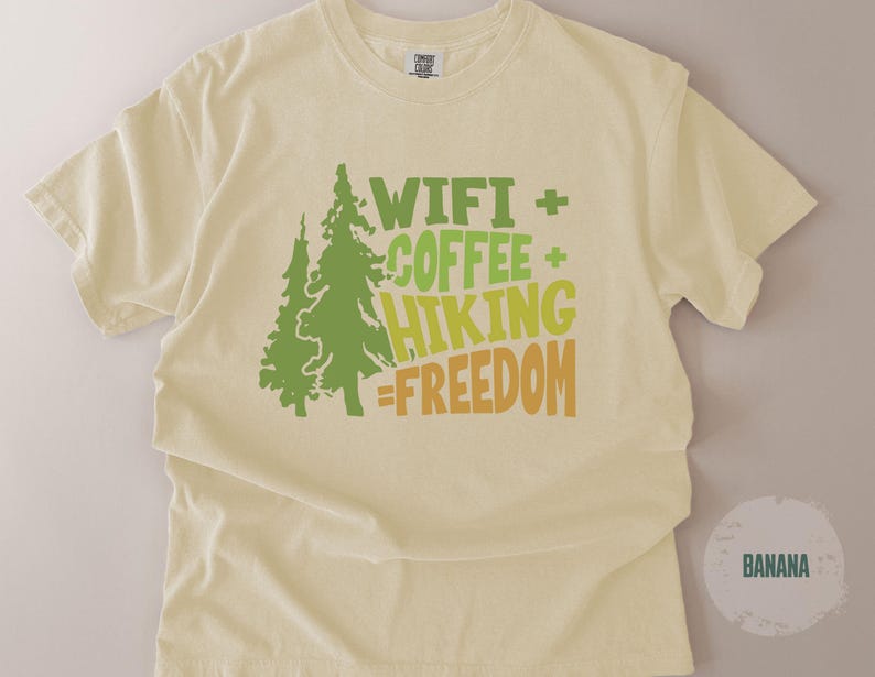 A light yellow t-shirt with the words "WIFI + COFFEE + HIKING = FREEDOM" in green and orange lettering. Two green tree graphics are on the left side of the shirt. The shirt is made of a soft material.