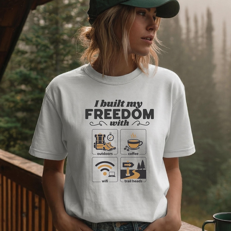 White t-shirt with the text "I built my FREEDOM with" and four icons: a backpack, a coffee cup, a wifi symbol, and a trail sign. The shirt is a crew neck and has short sleeves.