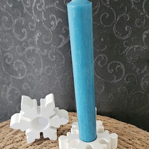 Può includere: Una candela alta, azzurra, è inserita in un portacandele bianco a forma di fiocco di neve. Accanto c'è un altro portacandele bianco a forma di fiocco di neve. La candela e i portacandele sono su una superficie tessuta, con uno sfondo a motivi.