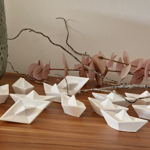Op de afbeelding: Een verzameling kleine, witte keramische papieren bootjes op een houten oppervlak. Decoratieve takken met roze bladeren en een textuurvaas op de achtergrond. De bootjes hebben een minimalistisch ontwerp.